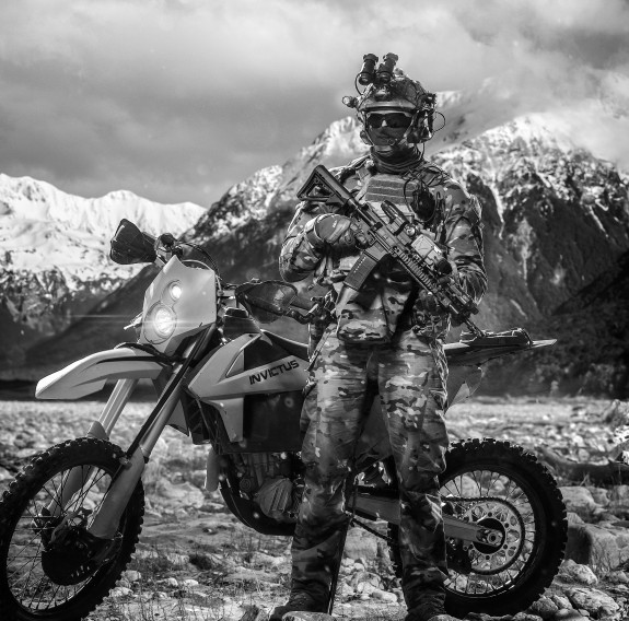 The image is black and white. A soldier stands in front of a snow-capped mountain range in military uniform. The soldier is wearing sunglasses, a helmet with a head set and night vision goggles as well as holding a weapon. The soldier is standing in front