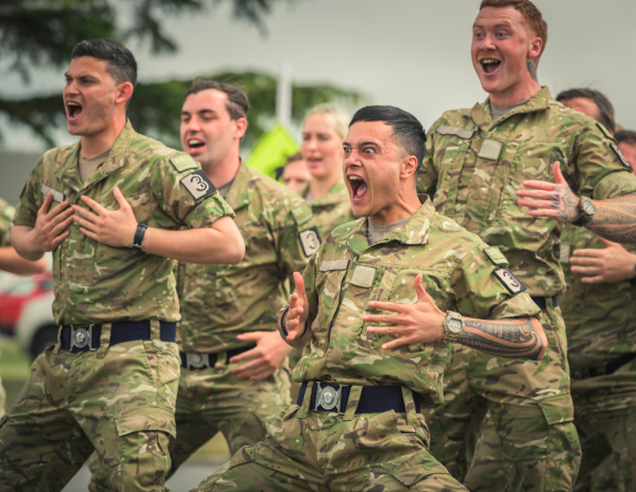 Soldiers in full uniform are pictured mid-haka, knees bent, hands on chest and mouths open.