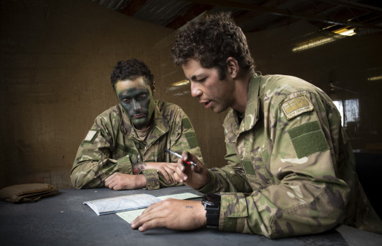 Lance Corporal Luca Soares Gonzalez sitting at a desk in the classroom, looking down at paperwork while his comrade also sits and looks on.
