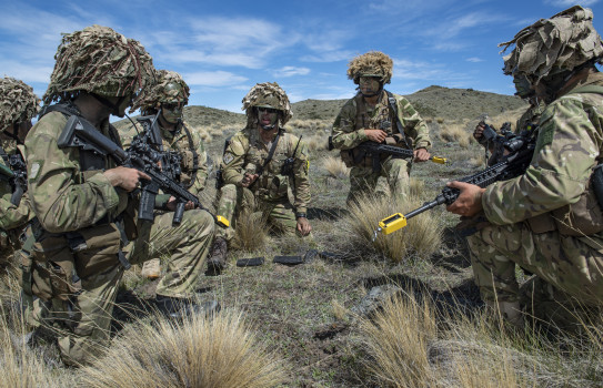 Seven soldiers in full operational gear group together in a circle to talk tactics at the Tekapo Military Training Area. Tussock filled hills and a blue sky can be seen in the background.