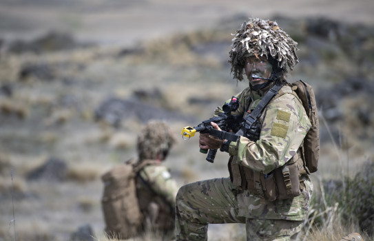 A soldier looks into the distance, taking a knee. They're holding a gun, and are flanked by another soldier in the distance.