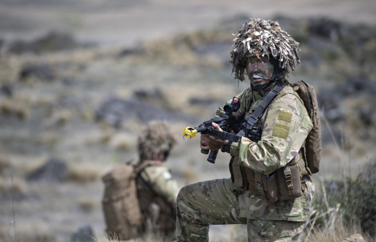 A soldier looks into the distance, taking a knee. They're holding a gun, and are flanked by another soldier in the distance.