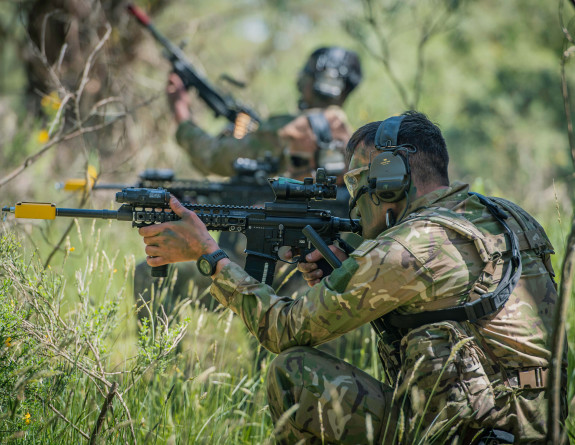 Soldiers are in a bushy and tree lined landscape, with face paint, earmuffs, and rifles. 