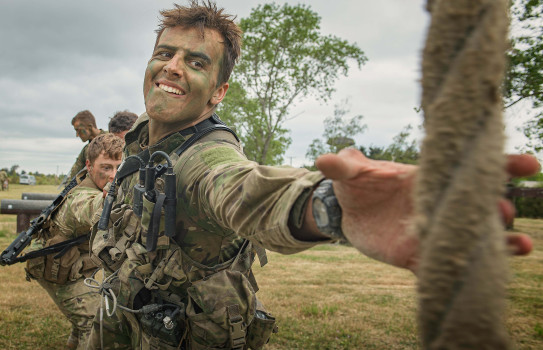 A line of soldiers link arms, as one reaches out to grab a rope.