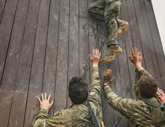 Two soldiers lift their comrade up and over a wall of an obstacle course