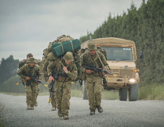 A group of uniformed soldiers with large packs march along a road. Faces are determined.