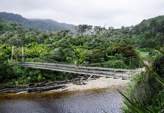 Two soldiers cross an elevated bridge, with a wide flowing river beneath and forest on either side.