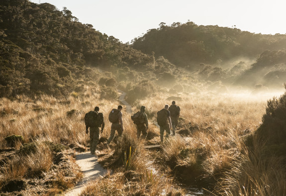 Five soldiers trek in single file, surrounded by a golden-brown tussock and hill landscape. Early morning sees steam rising as the day warms up.