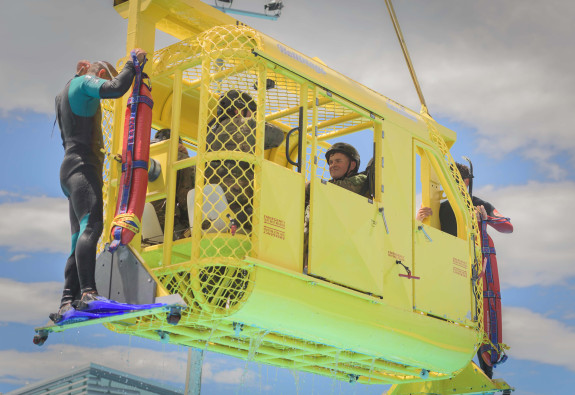 Soldiers in uniform and helmets are suspended in the air by a crane, while sitting in a small metal helicopter simulator 