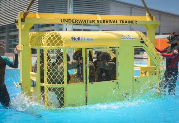 Soldiers in uniform and helmets are suspended in the air by a crane, while sitting in a small metal helicopter simulator 