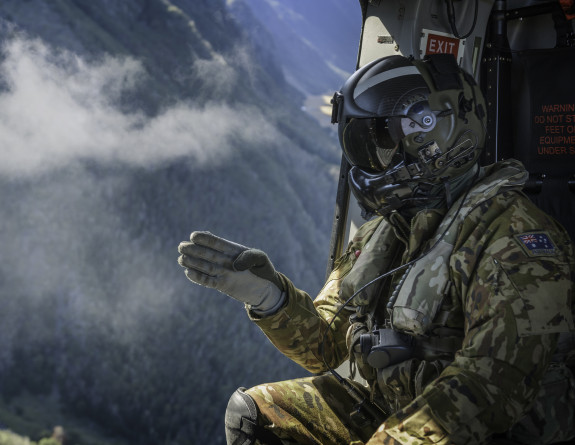 A Helicopter Loadmaster in a RNZAF helicopter points outside the aircraft. In the background you can see a mountainous range with some cloud cover. 