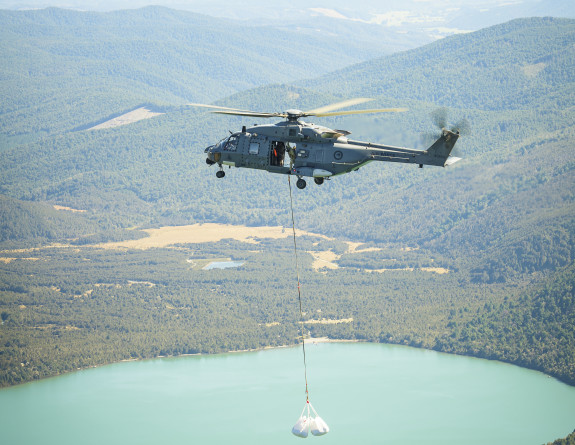 An NH90 helicopter carries an under slung load over the water. In the background there are mountain ranges. 
