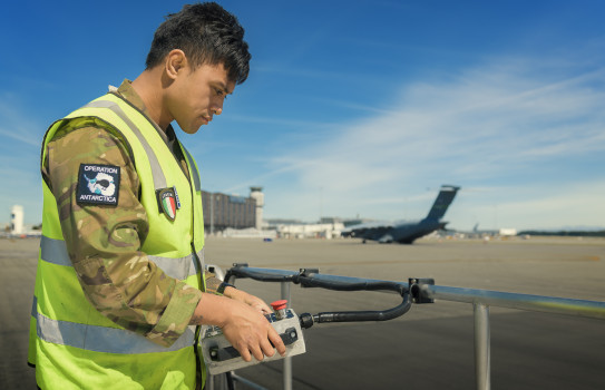 A member of the Antarctic program working in front of a Hercules aircraft on the tarmac under blue skies at the Harewood Terminal.