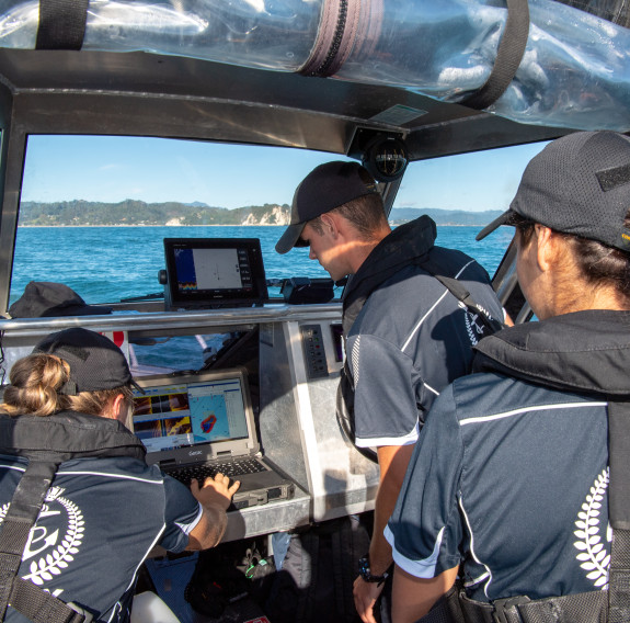 The team gather around the screens under the cover of the workboat’s cockpit on a sunny day.