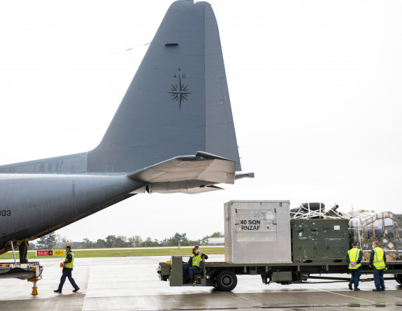 Royal New Zealand Air Force personnel prepare the Hercules aircraft, loading on supplies. The image shows the back of the aircraft with things being loaded via the ramp. 