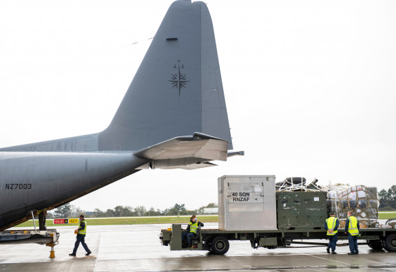 Royal New Zealand Air Force personnel prepare the Hercules aircraft, loading on supplies. The image shows the back of the aircraft with things being loaded via the ramp. 