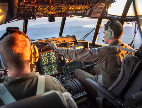 View from the Hercules aircraft flight deck as the crew transport military aid between staging centres in Europe. In the image there are two RNZAF personnel (pilot and co-pilot). There is sunlight in the flight deck. 