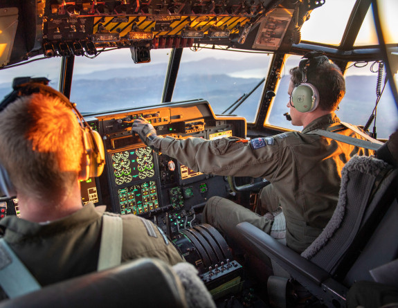 View from the Hercules aircraft flight deck as the crew transport military aid between staging centres in Europe. In the image there are two RNZAF personnel (pilot and co-pilot). There is sunlight in the flight deck. 