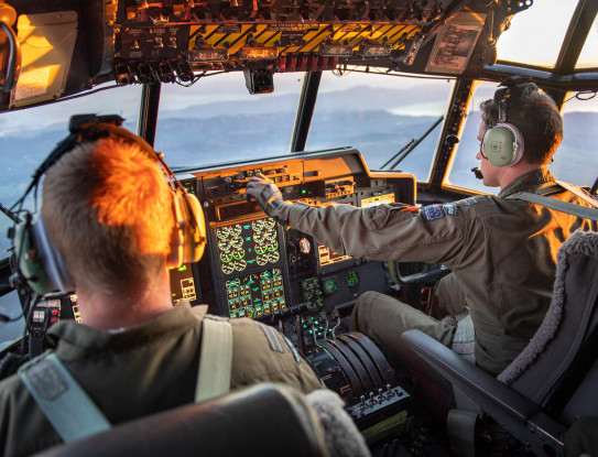 View from the Hercules aircraft flight deck as the crew transport military aid between staging centres in Europe. In the image there are two RNZAF personnel (pilot and co-pilot). There is sunlight in the flight deck.