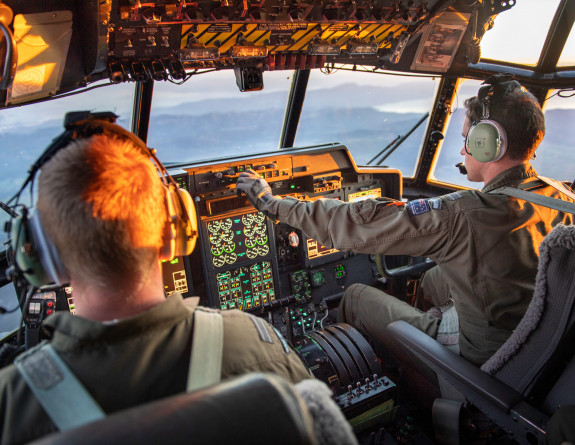 View from the Hercules aircraft flight deck as the crew transport military aid between staging centres in Europe. In the image there are two RNZAF personnel (pilot and co-pilot). There is sunlight in the flight deck. 