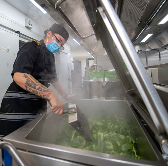 A chef is cooking in the kitchen of the Vince McGlone galley in preparation for service as the steam fogs the chef's glasses.