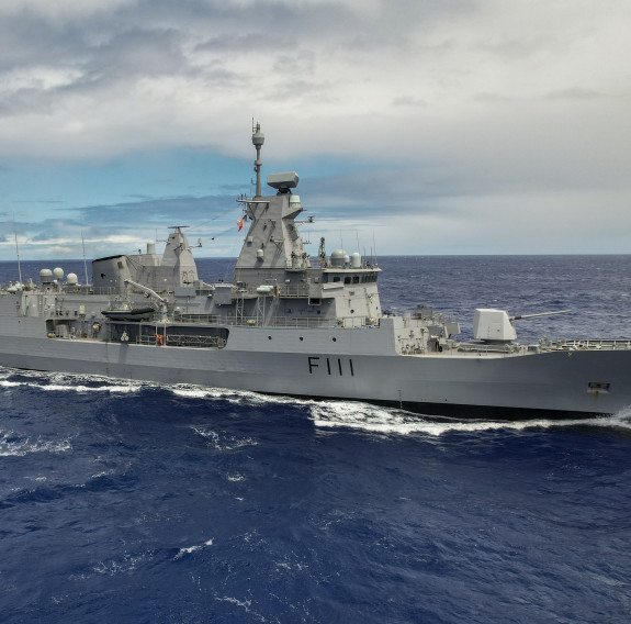 A stock photo of HMNZS Te Mana. A grey frigate cuts through the sea on an overcast day, the ship has a large 5" gun on the front of the ship..