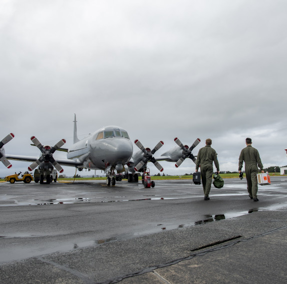 Two personnel walk towards the P-3K2 Orion on the tarmac at Base Auckland. The control tower is visible in the background on a wet and overcast day.