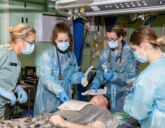 Four nurses work on a patient onboard a ship, they are all wearing blue scrubs/uniform, face masks and gloves. 