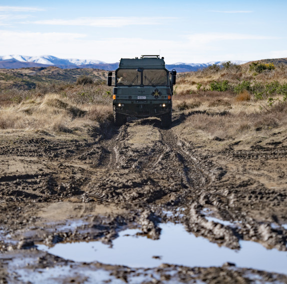 MHOV driving towards the camera down a mud track with a puddle in the foreground. Snow capped mountains can be seen in the background.