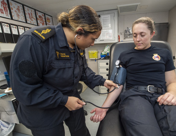 A woman wearing a dark blue Navy uniform with gold accents checks blood pressure of patient in a medical room.