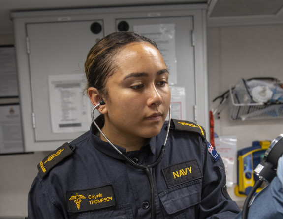 A woman wearing a dark blue Navy uniform with gold accents checks blood pressure of patient in a medical room.