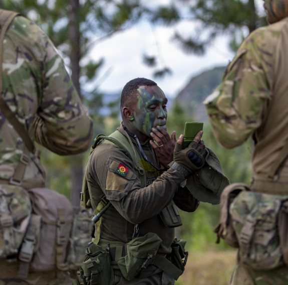 A Republic of Fiji Military Forces soldier puts camouflage paint on his face, two other soldier stand in the foreground. 