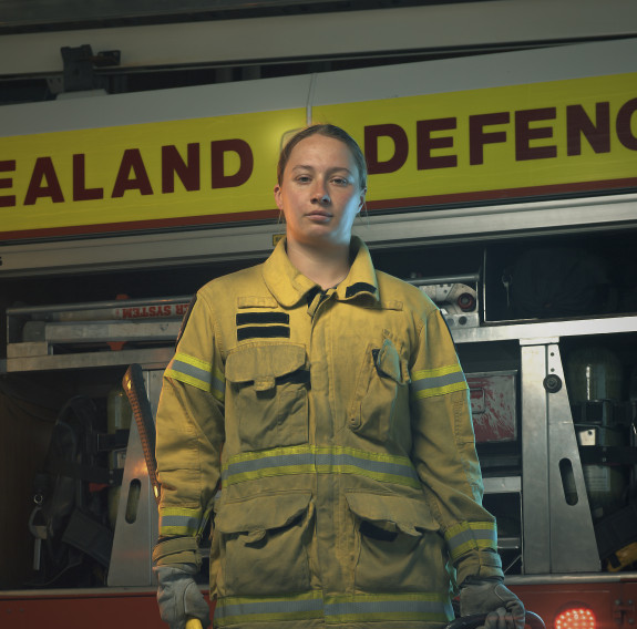 Sapper Danielle Brooks in a yellow firefighter jacket and black pants, looks and walks towards the camera while holding an axe in one hand, a jacket in the other with a red firetruck in the background