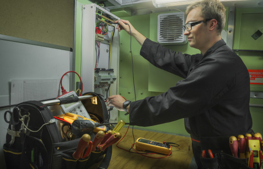 A man wearing dark overalls, safety classes and a tool belt works on an electrical box with tools in the foreground.