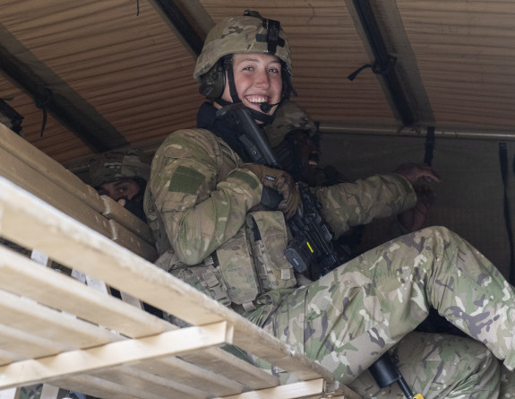 A soldier smiles as the camera while sitting elevated in the back of an Army vehicle.