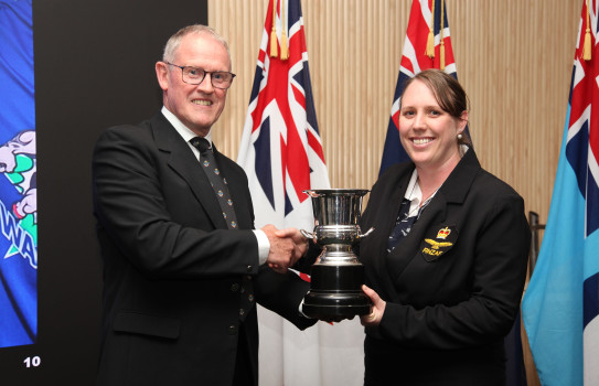 A man (left) shakes hand with a woman (right), they smile and shake hands as a trophy is presented in front of three flags.