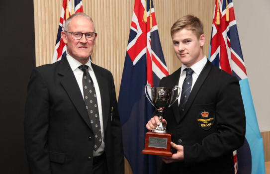 Two men wearing suits smile, one holds a trophy in front of three flags.