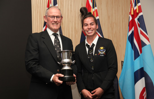 A man and a woman wearing suits smile, one holds a trophy in front of three flags.