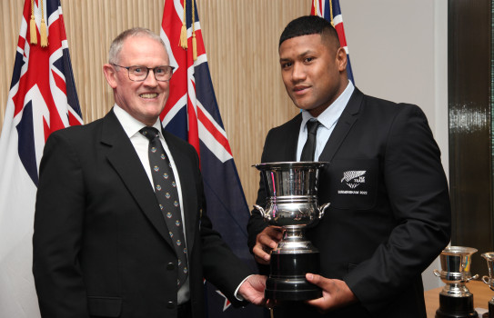 Two men wearing suits smile, one holds a trophy in front of three flags.