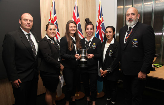 Six people wearing suits smile, two hold each side of a trophy in front of three flags.