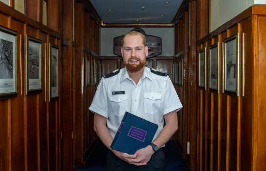 Midshipman James Olsen wearing Navy dress whites, stands in a wood panelled hallway holding a blue folder that reads, 'New Zealand Courts Martial Appeal Reports'.
