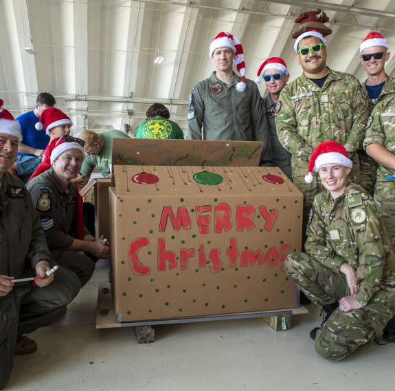 RNZAF personnel wearing a variety of uniforms and santa hats smile as they stand around a cardboard that reads "Merry Christmas".