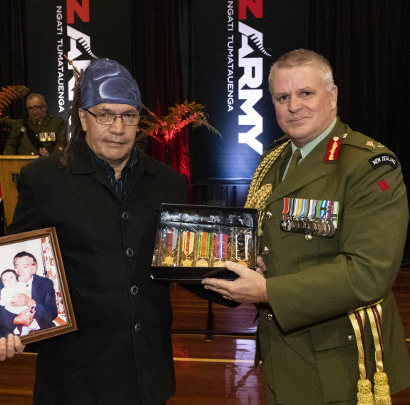 A man wearing a blue bandana and glasses receives medals from the Chief of Army, who is wearing a formal dark green uniform.