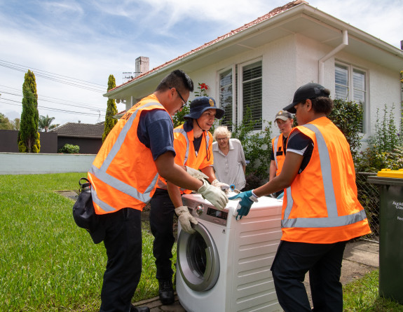 Navy personnel wearing high vis stand around a dryer that they have removed from a woman's home.