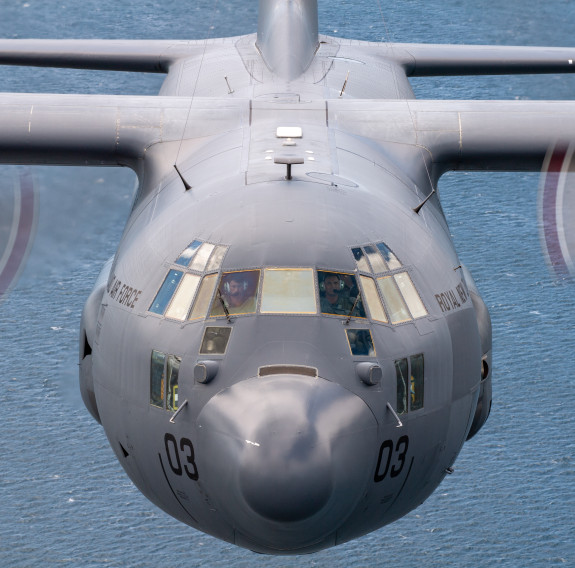 Nose-on air to air view of the Hercules aircraft, two pilots are visible in the cockpit and the number '03' is mirrored on the front of the aircraft, just below the windows.