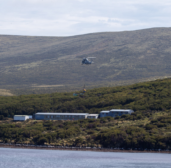 A Seasprite helicopter lifts cargo over buildings on Campbell Island, the ocean is visible in the foreground and cloud skies in the background.