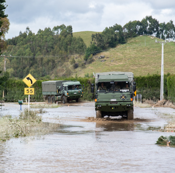 Two green Army trucks travel around a hedged corner into flood waters on the way to Moteo from Napier. A rounded hill with a tree line sits in the background and the water stretched to the foreground.