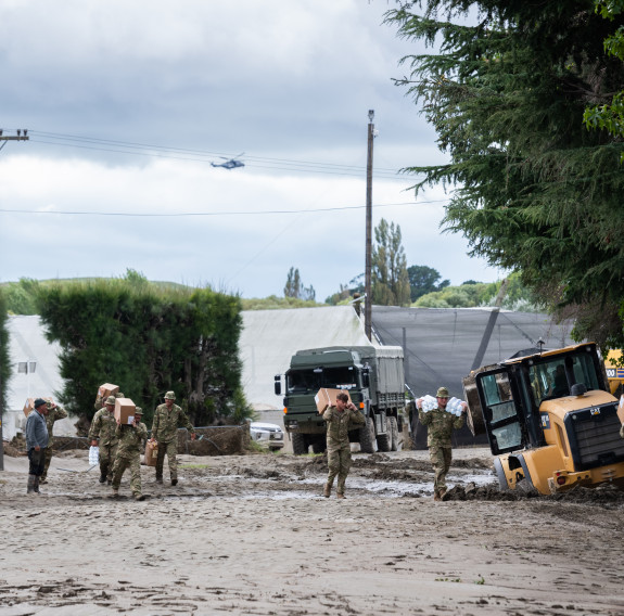 Soldiers carry boxes on their backs through mud to a local's ATV to transport goods to Moteo Marae. An NH90 helicopter appears in the background. A yellow digger is stuck in the mud on the right of the frame.