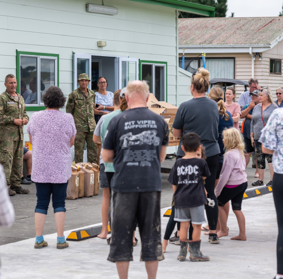 Soldiers talk with the locals who have congregated around them. They are standing in front of two buildings in a car park with jerry cans and supplies.