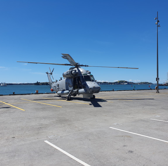 A wide shot of a grey Seasprite helicopter parked in a car park on wharf, next to the ocean on a sunny day.